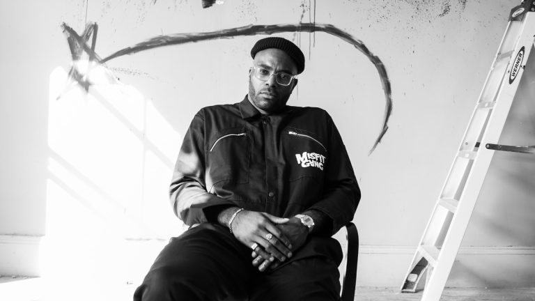 A black and white photos of Chiedu, sitting looking at the camera, in a white room with painting on the wall behind him.